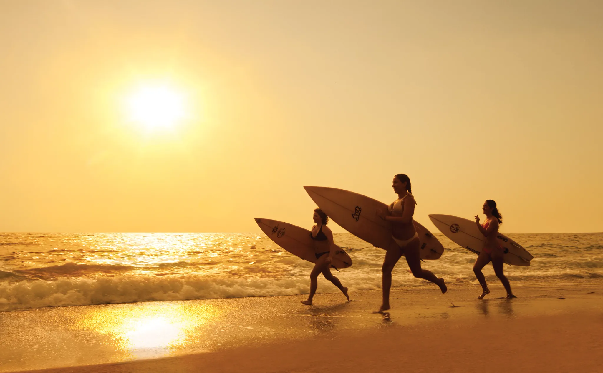 Women carrying surfboards along the beach at sunset near Dreams Vallarta Bay Resort & Spa.